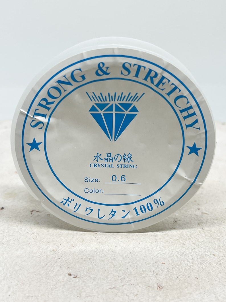 Puede incluir: Una bobina blanca de hilo de cristal fuerte y el&aacute;stico, etiquetada como "Strong & Stretchy" y "Crystal String". La bobina est&aacute; etiquetada con el tama&ntilde;o "0,6" y el material "Poliuretano 100&nbsp;%".