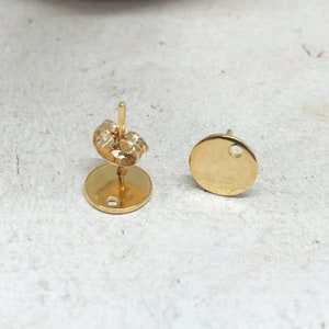 May include: A pair of gold-toned earring blanks with a round shape and a small hole in the center. The earring backs are also gold-toned.