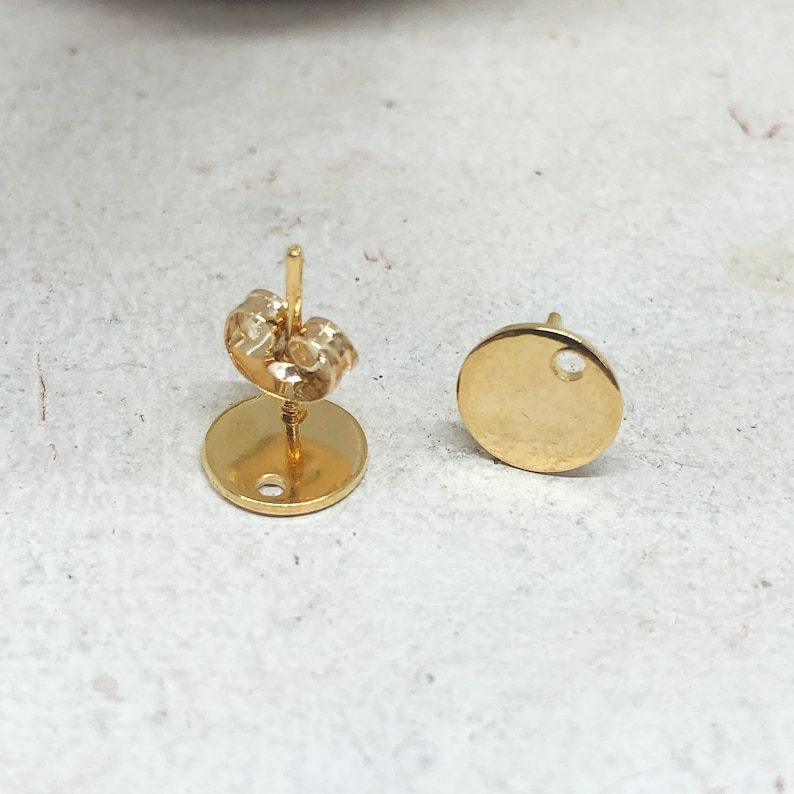 May include: A pair of gold-toned earring blanks with a round shape and a small hole in the center. The earring backs are also gold-toned.