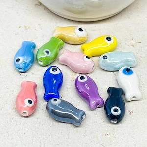 Könnte beinhalten: Eine Sammlung von zwölf kleinen, glatten, fischförmigen Glasperlen in verschiedenen Farben wie Blau, Grün, Gelb, Rosa, Violett, Weiß und Dunkelgrau. Jede Perle weist ein einzelnes, markantes Augendetail mit schwarzer Pupille und weißer Iris auf.