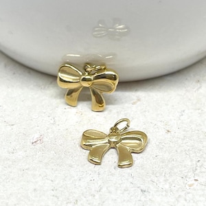 Op de afbeelding: Twee kleine, gepolijste goudkleurige metalen bedels in de vorm van strikjes. Elke bedel heeft een glad, reflecterend oppervlak en een klein rond ringetje aan de bovenkant in het midden, waardoor ze gemakkelijk aan kettingen kunnen worden bevestigd.