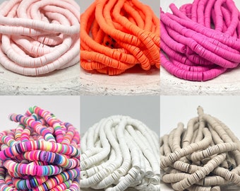 May include: Assortment of colorful clay beads in various colors, including pink, orange, white, beige, and multi-colored. The beads are arranged in piles on a white surface.