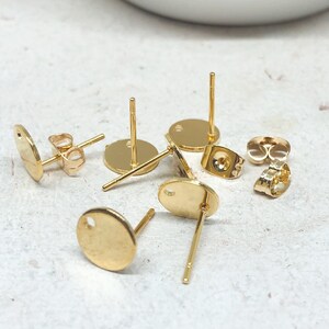 May include: Gold-toned earring blanks with a round shape and a hole in the center. The earring blanks are shown on a light gray surface.