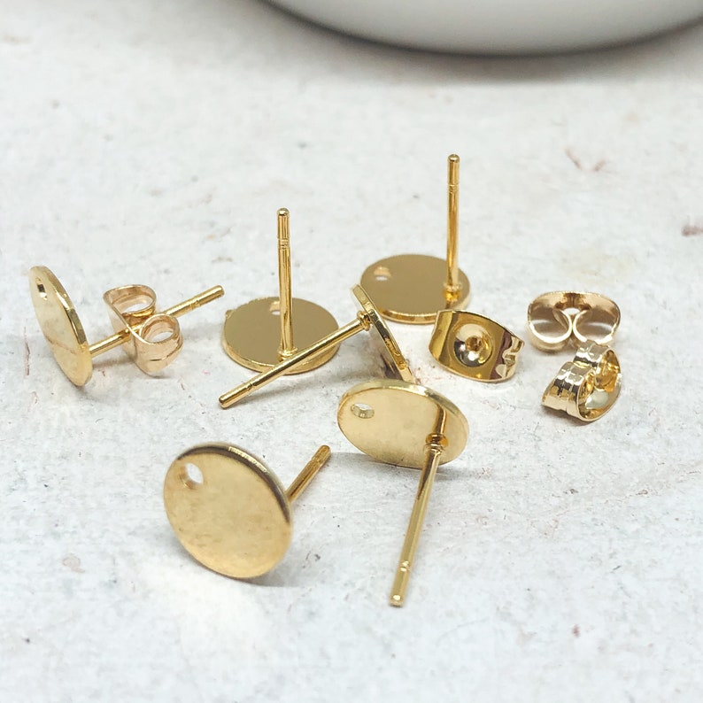 May include: Gold-toned earring blanks with a round shape and a hole in the center. The earring blanks are shown on a light gray surface.