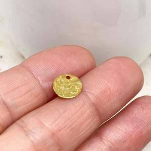 May include: A small, circular, gold-coloured charm with a hammered texture and a small hole at the top. The charm is held between two fingers, highlighting its size and detail. The charm is likely used for jewellery making or other craft projects.