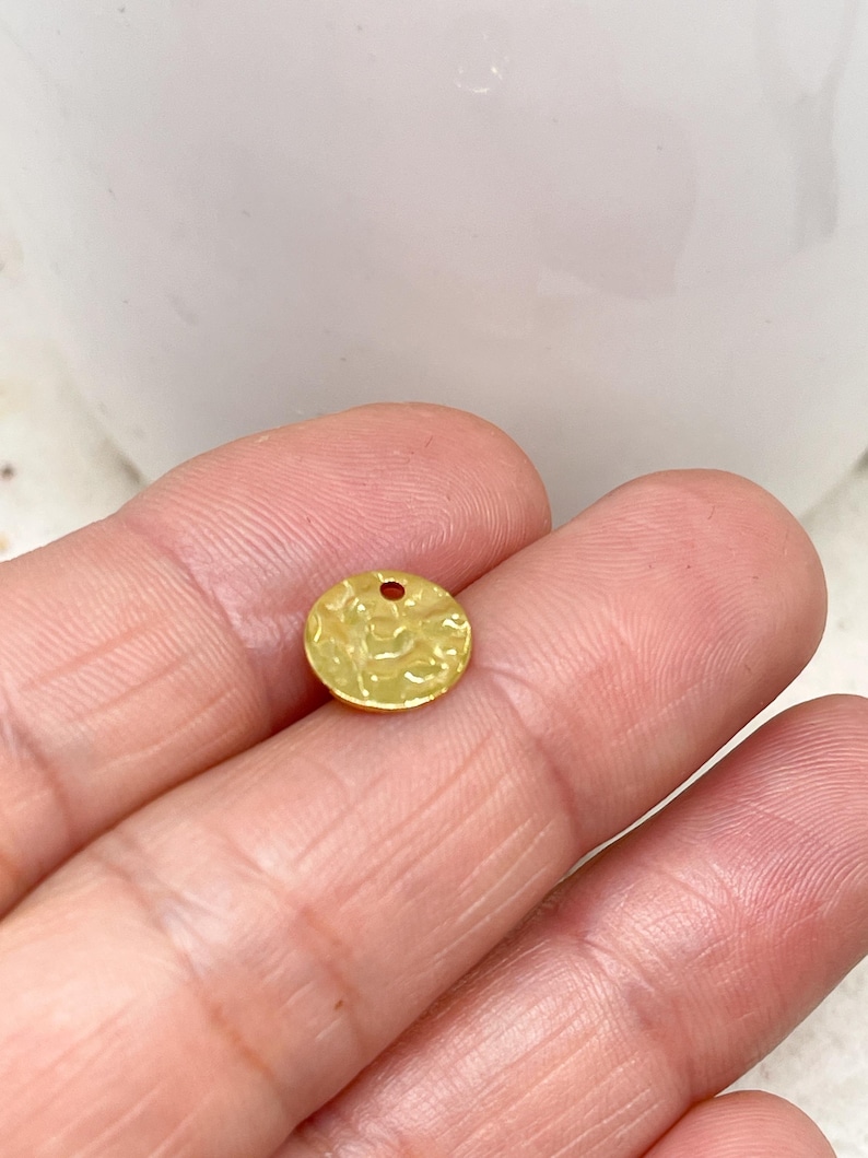 May include: A small, circular, gold-coloured charm with a hammered texture and a small hole at the top. The charm is held between two fingers, highlighting its size and detail. The charm is likely used for jewellery making or other craft projects.