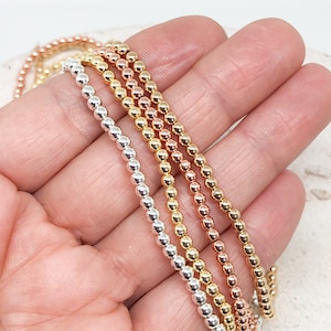Könnte beinhalten: Nahaufnahme mehrerer Perlenketten in Silber, Gold, Roségold und Gelbgold. Die Halsketten bestehen aus kleinen, runden Perlen, die aneinandergereiht sind. Die Perlen haben eine glänzende, metallische Oberfläche. Die Halsketten werden auf hellem Hintergrund präsentiert.
