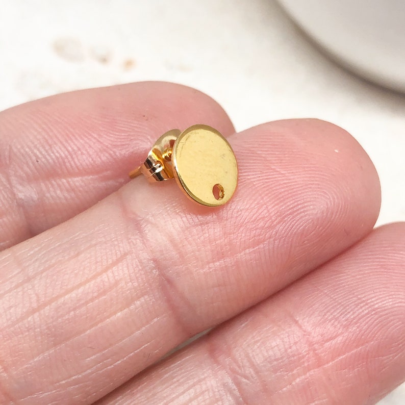 May include: Gold-toned earring with a round disc and a small hole in the center. The earring is on a white background.