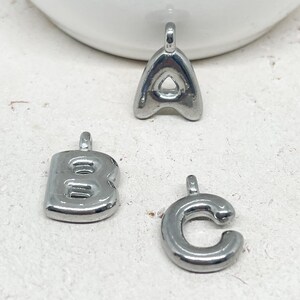 May include: Three silver-colored metal alphabet charms: the letters A, B, and C. Each charm has a small loop at the top for attaching to a chain or bracelet. The letters have a rounded, bubble-like design.
