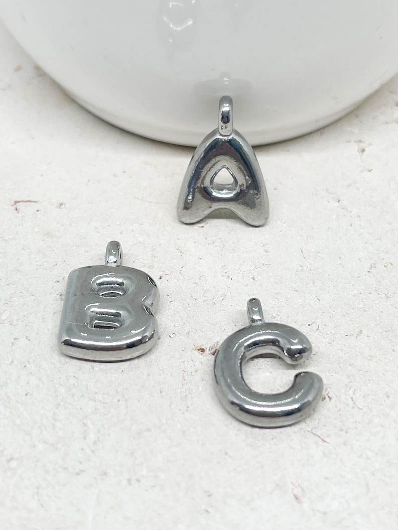 May include: Three silver-colored metal alphabet charms: the letters A, B, and C. Each charm has a small loop at the top for attaching to a chain or bracelet. The letters have a rounded, bubble-like design.