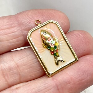 May include: A rectangular gold-toned pendant featuring a colorful fish design. The fish has a gold head, red, green, and yellow scales, and a black tail. The pendant has a cream-colored background with a pinkish glow and a gold border.