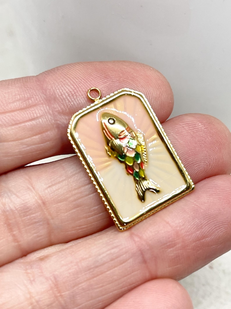 May include: A rectangular gold-toned pendant featuring a colorful fish design. The fish has a gold head, red, green, and yellow scales, and a black tail. The pendant has a cream-colored background with a pinkish glow and a gold border.