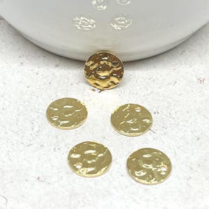 May include: Five gold-coloured, hammered-finish metal disc charms. Each charm has a slightly irregular, textured surface and a small hole for stringing. The charms are round and vary slightly in size. The charms are for jewellery making.