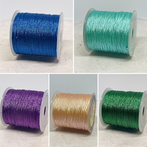 Könnte beinhalten: Vier Spulen mit Metallic-Garn in verschiedenen Farben: Türkis, Lila, Gold und Grün. Die Spulen sind alle gleich groß und haben ein transparentes Kunststoffgehäuse.