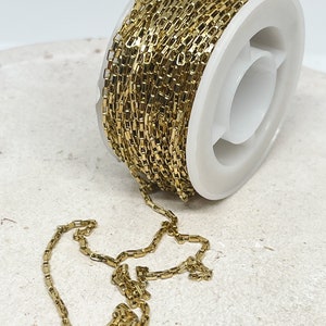 Könnte beinhalten: Eine Spule mit goldfarbener Kette mit rechteckigem Gliederdesign. Die Kette ist um eine weiße Kunststoffspule gewickelt. Ein Teil der Kette ist abgewickelt und liegt auf einer strukturierten, cremefarbenen Oberfläche.