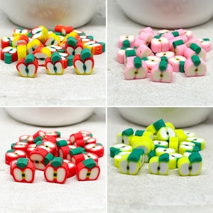 Könnte beinhalten: Sortiment an Polymer Clay-Apfelperlen in den Farben Rot, Gelb, Rosa und Grün. Die Perlen haben ein grünes Blattdetail und sind perfekt für die Schmuckherstellung oder zum Basteln.