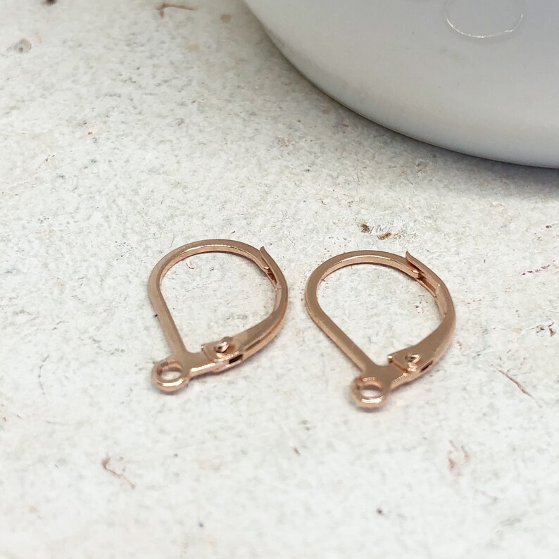 May include: Two rose gold earring hooks, also known as French hooks, are shown on a light gray surface. The hooks are made of metal and have a smooth finish.