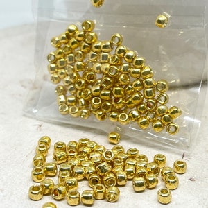 May include: A close-up of a pile of gold-colored metal beads. The beads are small and round, with a smooth, shiny finish. They are scattered on a white surface.