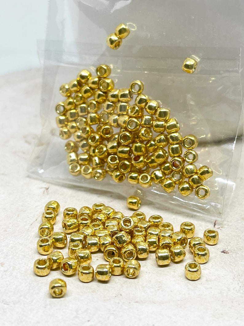 May include: A close-up of a pile of gold-colored metal beads. The beads are small and round, with a smooth, shiny finish. They are scattered on a white surface.
