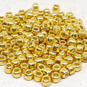 May include: A pile of gold-colored seed beads. The beads are small and round, and they have a shiny finish.