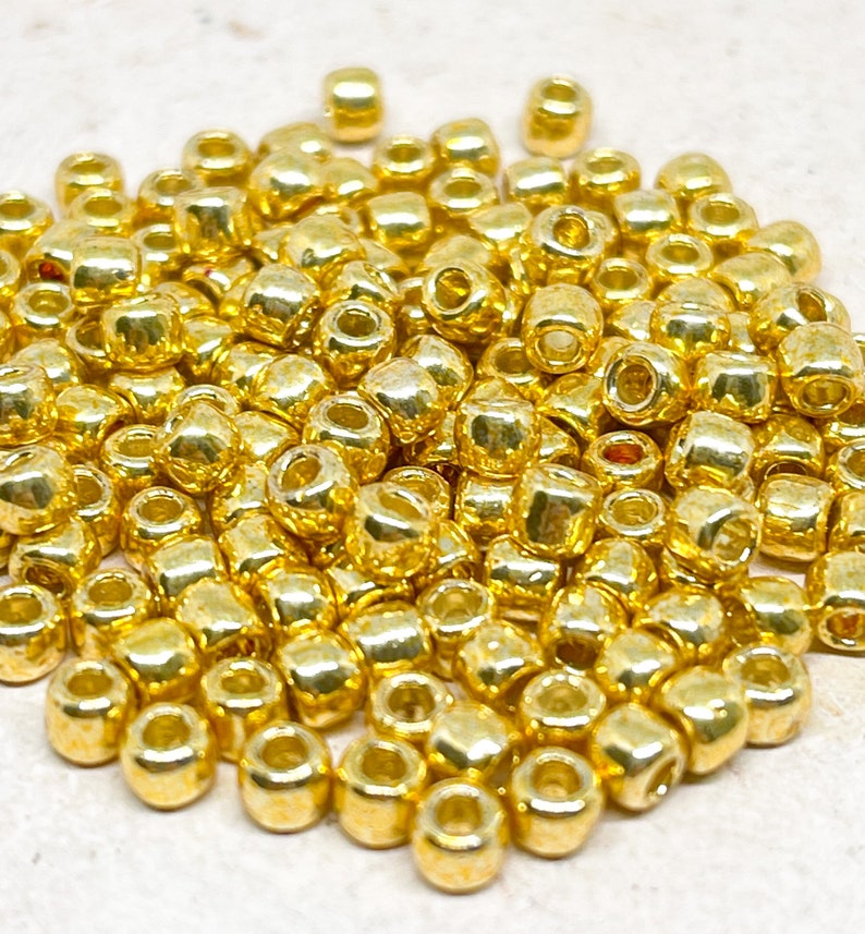 May include: A pile of gold-colored seed beads. The beads are small and round, and they have a shiny finish.
