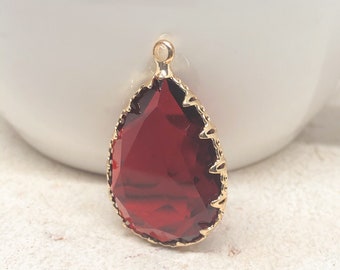 May include: A teardrop-shaped pendant featuring a deep red faceted gemstone set within a gold-toned metal frame. The setting has a decorative scalloped edge and a small circular loop at the top for attachment, showcasing a polished finish.
