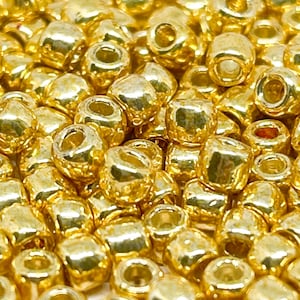 May include: A close-up image of a pile of gold-colored seed beads. The beads are round and have a shiny finish.