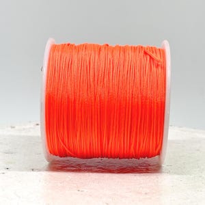 Könnte beinhalten: Eine Spule mit leuchtend neonorangefarbener Nylonschnur, die fest um einen weißen Kunststoffzylinder gewickelt ist. Die Schnur ist dünn und glatt und erzeugt eine gleichmäßige Textur auf der gesamten Oberfläche der aufrecht stehenden Spule.