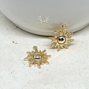 Op de afbeelding: Twee goudkleurige zonbedels met gedetailleerde stralen en een centraal cirkelvormig ontwerp. De bedels hebben een lus aan de bovenkant om te bevestigen. Lichte achtergrond.