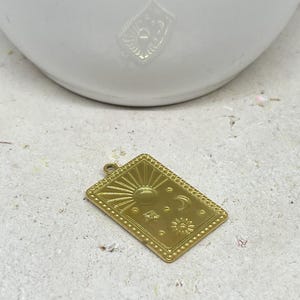 Puede incluir: Un colgante rectangular dorado con dise&ntilde;os celestiales. El colgante tiene un sol, una luna, estrellas y un borde punteado. Se muestra sobre una superficie clara y texturizada, con un objeto blanco en el fondo.