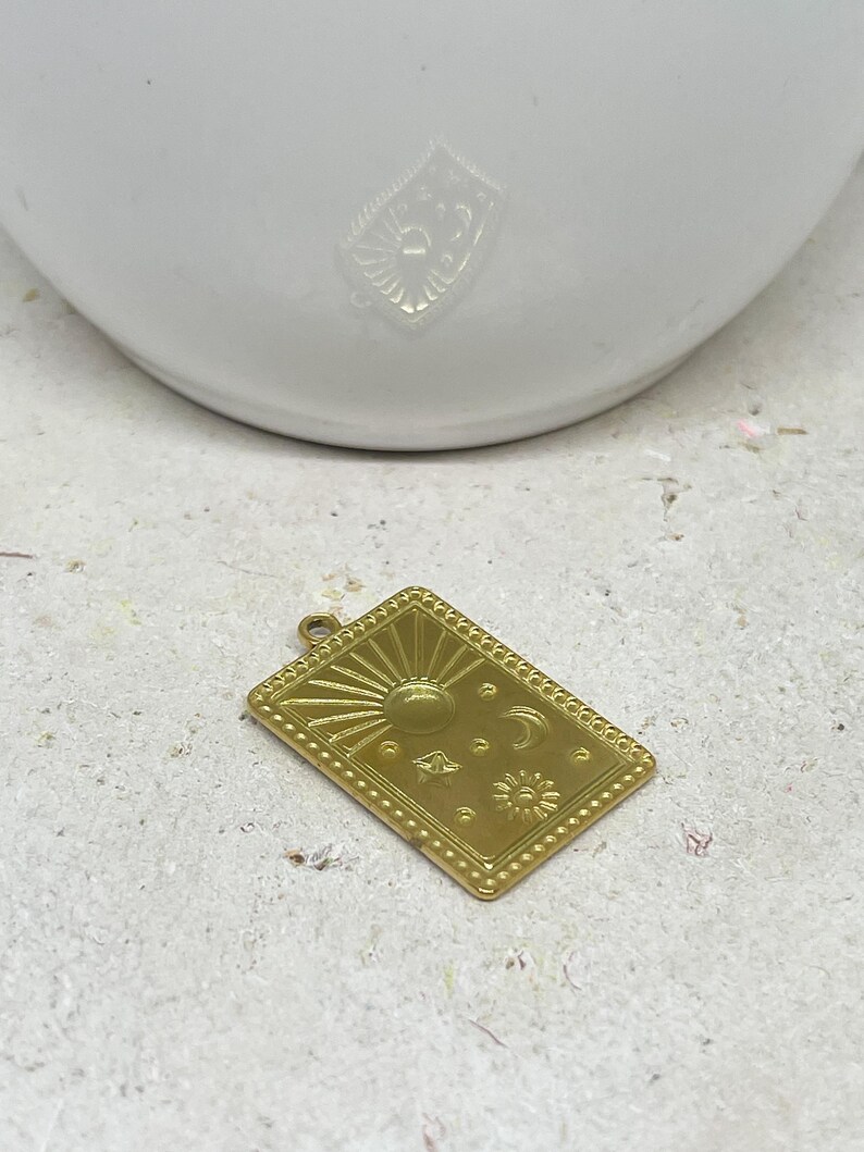 Puede incluir: Un colgante rectangular dorado con dise&ntilde;os celestiales. El colgante tiene un sol, una luna, estrellas y un borde punteado. Se muestra sobre una superficie clara y texturizada, con un objeto blanco en el fondo.