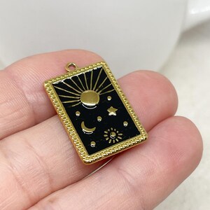 Puede incluir: Un dije rectangular dorado con una incrustaci&oacute;n de esmalte negro que presenta s&iacute;mbolos celestiales: un sol, una luna, estrellas y un sol m&aacute;s peque&ntilde;o. El dije tiene un borde decorativo y un peque&ntilde;o lazo para sujetar, ideal para hacer joyas.