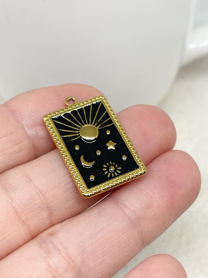 Puede incluir: Un dije rectangular dorado con una incrustaci&oacute;n de esmalte negro que presenta s&iacute;mbolos celestiales: un sol, una luna, estrellas y un sol m&aacute;s peque&ntilde;o. El dije tiene un borde decorativo y un peque&ntilde;o lazo para sujetar, ideal para hacer joyas.