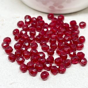 Könnte beinhalten: Eine Ansammlung kleiner, facettierter, tiefroter Glasperlen. Jede Perle weist eine mehrseitige, reflektierende Oberfläche auf, die das Licht einfängt und ein funkelndes Aussehen erzeugt. Die Perlen sind rund und haben eine einheitliche karmesinrote Farbe.