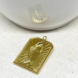 May include: A gold-colored rectangular pendant featuring a detailed fish design. The fish is centered within a frame, with radiating lines emanating from behind it. The pendant has a small loop at the top for attaching to a chain or other jewelry.
