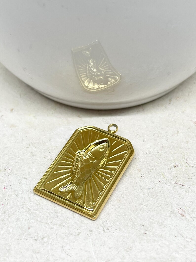 May include: A gold-colored rectangular pendant featuring a detailed fish design. The fish is centered within a frame, with radiating lines emanating from behind it. The pendant has a small loop at the top for attaching to a chain or other jewelry.
