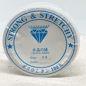Puede incluir: Una bobina blanca de hilo de cristal fuerte y el&aacute;stico, etiquetada con "Strong & Stretchy" y "Crystal String". La bobina est&aacute; etiquetada con el tama&ntilde;o "0,8" y el texto "Water's Line". La bobina tambi&eacute;n est&aacute; etiquetada con el texto "Poliuretano 100%".