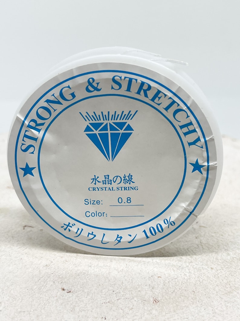 Puede incluir: Una bobina blanca de hilo de cristal fuerte y el&aacute;stico, etiquetada con "Strong & Stretchy" y "Crystal String". La bobina est&aacute; etiquetada con el tama&ntilde;o "0,8" y el texto "Water's Line". La bobina tambi&eacute;n est&aacute; etiquetada con el texto "Poliuretano 100%".