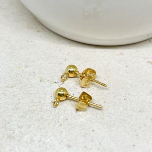 May include: Two gold-colored stud earrings with ball-shaped fronts and post backs. The earrings are displayed on a light-colored, textured surface. The earrings are small and delicate.