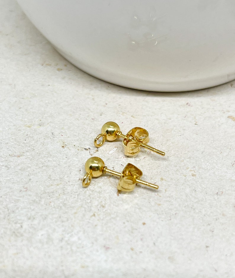 May include: Two gold-colored stud earrings with ball-shaped fronts and post backs. The earrings are displayed on a light-colored, textured surface. The earrings are small and delicate.