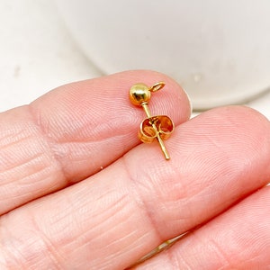 May include: A single gold-colored earring post with a small ball at the top and a butterfly clutch. The earring post is held in a hand, showing the details of the jewelry piece. The earring post is a simple, elegant design.