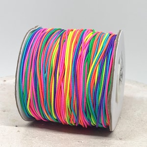 Könnte beinhalten: Eine Spule mit bunten Gummibändern in Regenbogenfarben, darunter Pink, Orange, Gelb, Grün, Blau und Lila. Das Band ist eng auf eine Pappspule gewickelt, ideal für Bastelarbeiten und Schmuckherstellung.
