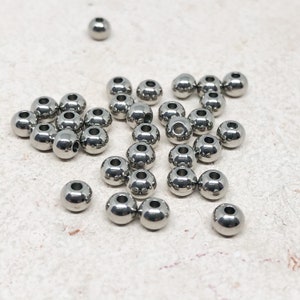 May include: A collection of silver metal beads, scattered on a light gray surface. The beads are round and have a smooth, shiny finish.