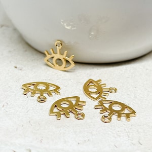 Könnte beinhalten: Fünf goldfarbene Augen-Anhänger mit einer kleinen Schlaufe oben. Die Augen sind mit einer dickeren Linie umrandet und haben Wimpern.