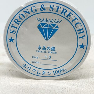 Puede incluir: Carrete blanco de hilo de cristal fuerte y el&aacute;stico, etiquetado como "Strong & Stretchy" y "Crystal String". El carrete tambi&eacute;n tiene el texto "Size: 1.0" y "Poliuretano 100%".
