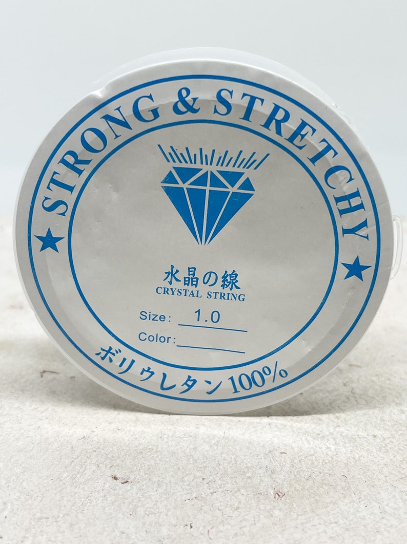 Puede incluir: Carrete blanco de hilo de cristal fuerte y el&aacute;stico, etiquetado como "Strong & Stretchy" y "Crystal String". El carrete tambi&eacute;n tiene el texto "Size: 1.0" y "Poliuretano 100%".