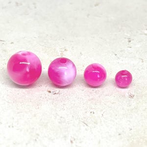 Könnte beinhalten: Vier kugelförmige Perlen, angeordnet in einer Reihe von der größten zur kleinsten. Jede Perle hat eine leuchtend pinke Farbe mit wirbelnden weißen Marmormustern und ein zentrales Loch zum Auffädeln. Die Perlen haben eine glatte Oberfläche.