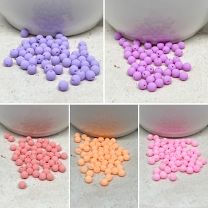 Könnte beinhalten: Eine Sammlung von pastellfarbenen runden Perlen in Lavendel, Pfirsich und Rosa. Die Perlen sind auf einer weißen Oberfläche verstreut.