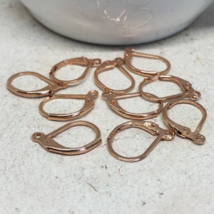 May include: Ten rose gold earring hooks, also known as ear wires, are scattered on a white surface. The hooks are made of metal and have a smooth finish.