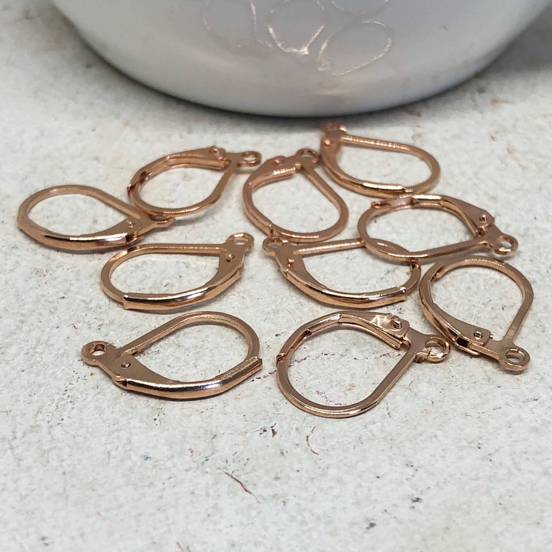 May include: Ten rose gold earring hooks, also known as ear wires, are scattered on a white surface. The hooks are made of metal and have a smooth finish.